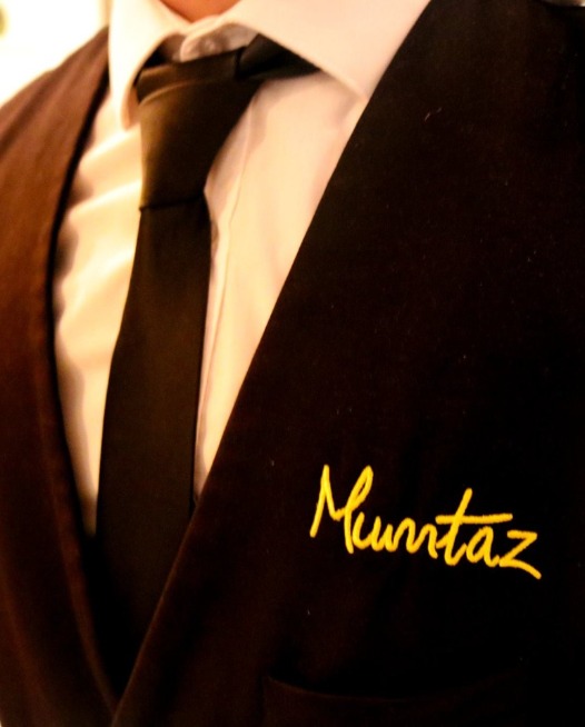 Close up of a waiter dressed in white shirt with tie and black jacket with 'Mumtaz' written on his top jacket pocket